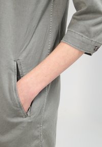 Vaaleanharmaa vaate, joka on rentoa mallia, siinä on näkyvä sivutasku ja käännetty hihansuu napin yksityiskohdalla. Pehmeä, sileä pinta.