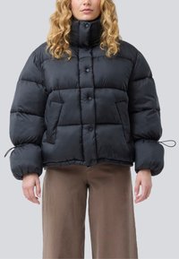 Žena v černé péřové bundě s vysokým límcem, zapínáním na knoflíky a šňůrkovými manžetami, v kombinaci s volnými hnědými kalhotami na jednoduchém pozadí.