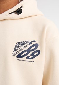 Sudadera crema con un acabado texturizado, que presenta un logotipo gráfico azul "NORTHWOOD PETROL IND. SINCE 89" y "GREEN GOLD LUMBERCREW."
