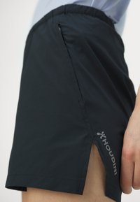 Svarte treningsshorts laget av lett materiale, med en sidelomme med glidelås, en spalte-design og en logo nær kanten.