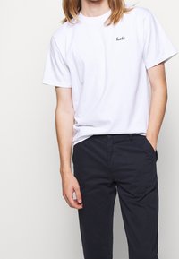 Witte katoenen T-shirt met korte mouwen en een zwart logo "forét" op de linkerborst, gecombineerd met marineblauwe broek. Minimalistisch ontwerp.