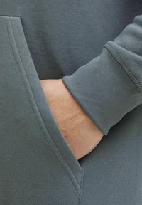 Grå sweatshirt med en blød tekstur, der har en frontlomme og manchetter ved ærmerne. Synlige syninger langs sømmene.