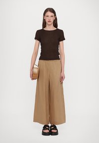 Hnědý strukturovaný fitted top, široké béžové kalhoty a černé platformové sandály. Nese malou pruhovanou dřevěnou kabelku. Jednoduchý design, minimální detaily.