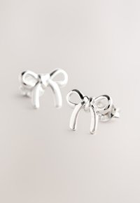 Srebrne naušnice u obliku mašnica, s glatkim linijama i poliranim površinama. Imaju jednostavan dizajn s kopčom na klipsu.