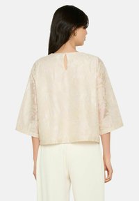 Blusa beige chiaro con motivo in pizzo floreale, maniche a tre quarti e dettaglio a chiave sul retro. Texture liscia e silhouette squadrata.