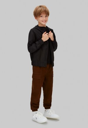 Czarna kurtka na zamek błyskawiczny z elastycznymi mankietami, w połączeniu z brązowymi spodniami typu cargo z kieszeniami i elastycznymi zakończeniami nogawek. Białe trampki z ozdobnymi logo.