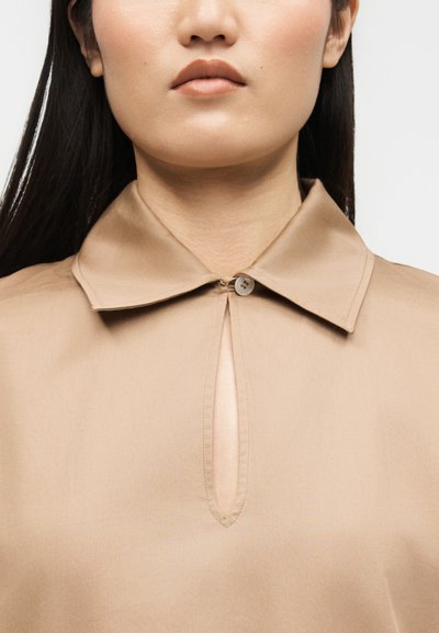Femme portant une chemise beige avec un col pointu fermé par un seul bouton au-dessus d'une ouverture verticale en forme de serrure.