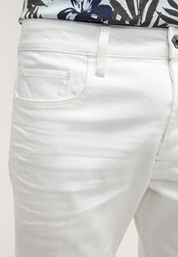 Jeans en denim blanc avec une texture lisse, un design classique à cinq poches et des détails de couture visibles. L'avant présente un zip et une fermeture à bouton.