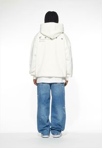 Felpa bianca oversize con cappuccio, con testo nero sul retro. Abbinata a jeans larghi strappati di colore blu chiaro e sneakers bianche.