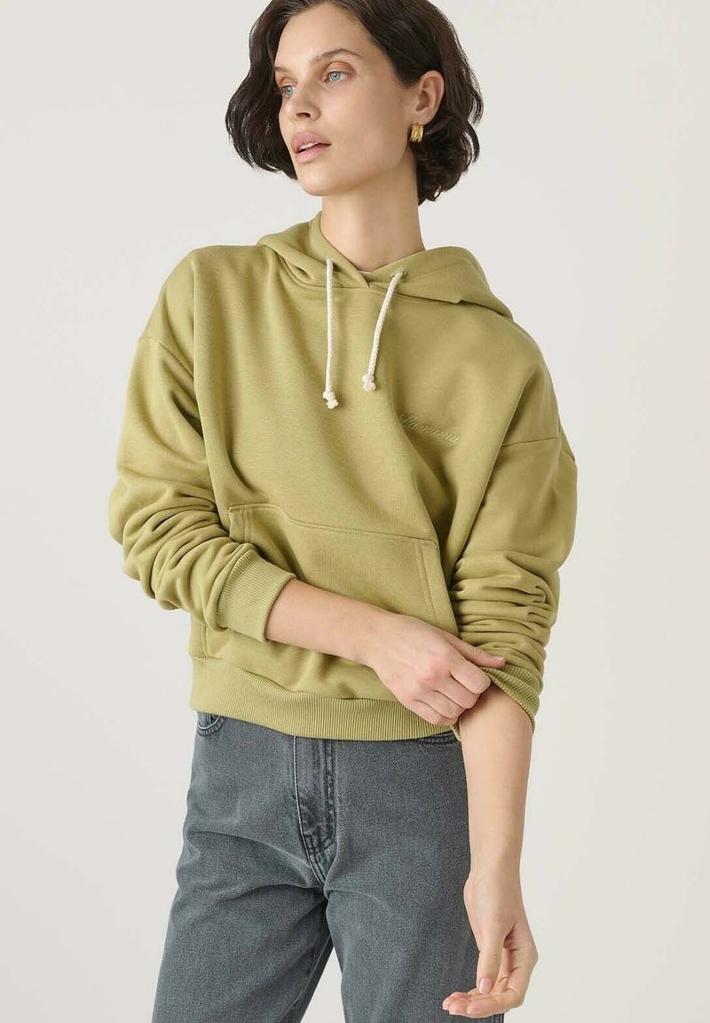 Olajzöld kapucnis felső, zsinórral, elülső zsebbel, bordás mandzsettákkal és aljjal, puha anyagból készült, szürke farmerral viselve.