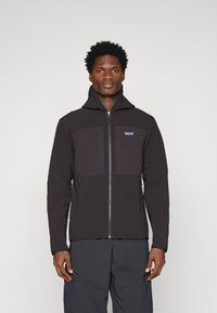 Juodas zip-up švarkas su tekstūruotu audiniu ir lygiomis, kontrastingomis detalėmis ant krūtinės, aukštu apykakle ir užtrauktukais šonuose.