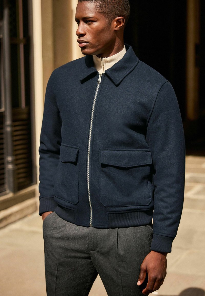 Marineblaue Bomberjacke aus weichem Material, mit einem Reißverschluss vorne und zwei großen Taschen. Getragen mit grauen, maßgeschneiderten Hosen.
