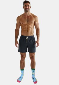 Mørkeblå badebukser til mænd med snørelukning i taljen, kombineret med farverige sokker med frugtmønstre på en lyseblå baggrund.