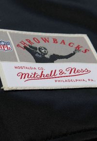 Mitchell & Ness Stoffetikett mit dem Text "Throwbacks", in Rot- und Grautönen, auf einem schwarzen Hintergrund genäht, versehen mit einer nostalgischen Illustration.