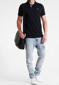 Černé polo tričko se šedým límcem, opotřebované světle modré džíny, bílé tenisky a černá taška přes rameno, to vše nosí mužská postava.