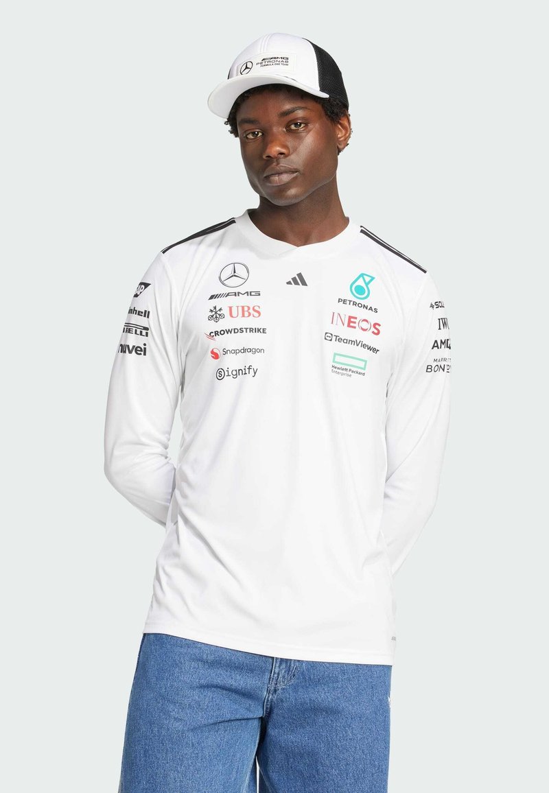 Camisa blanca de manga larga con detalles en negro, que presenta varios logotipos de patrocinadores. El modelo lleva una gorra en blanco y negro con un logotipo. Jeans azules.