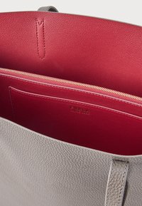 Světlá šedá kožená taška s otevřeným horním dílem, který odhaluje červený vnitřek, kapsu na zip a vyražené jméno značky na vnitřní kapse.