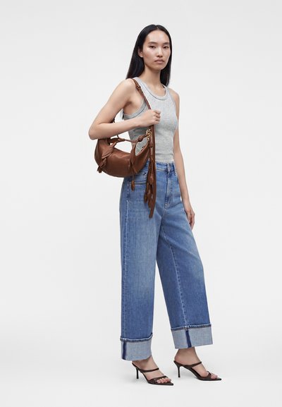 Femme portant un débardeur gris, un jean bleu large avec revers, des sandales noires à talons, et portant un sac à main en cuir marron avec des détails à franges.