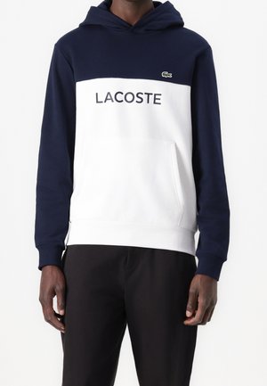 Man draagt een marineblauwe en witte Lacoste hoodie met logo en zwarte broek, staand tegen een effen witte achtergrond.