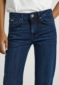 Jeans de denim azul oscuro con un diseño de cintura alta, bolsillos frontales y cremallera. Detalles de costura notables y una textura suave.