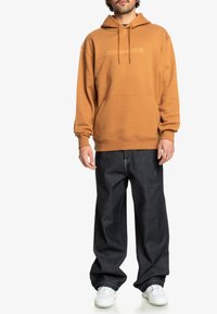 Bruine hoodie met een voorzak, voorzien van het 'DC Shoes'-logo in tonale tekst. Gemaakt in combinatie met losvallende donkere denim jeans en witte sneakers.
