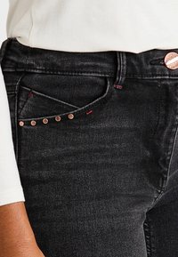 Primer plano de un bolsillo de unos jeans de mezclilla negros con remaches de cobre y una camisa blanca de manga larga parcialmente visible en el lado izquierdo.