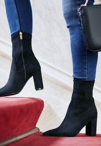 Schwarze Wildleder-Ankle Boots mit einem enganliegenden, sockenähnlichen Design, spitzem Fuß und einem klobigen Absatz, getragen mit Jeans auf einer rotteppichbelegten Treppe.