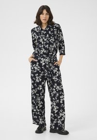 Mono floral en azul marino oscuro con flores blancas, mangas largas, cierre de botones en la parte delantera, cintura entallada y corte de pierna ancha. Zapatos negros completan el conjunto.