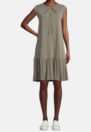 Kobieta nosząca bezrękawnikową, oliwkowozieloną sukienkę do kolan z falbaną u dołu i wiązaniem przy dekolcie, zestawioną z białymi trampkami.
