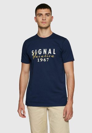 Fiatal férfi tengerészkék pólóban, melyen a "Signal Vacation 1967" felirat szerepel, bézs rövidnadrágban, áll egy egyszínű világos háttér előtt.