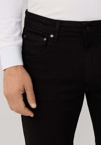 Jeans de mezclilla negros con un ajuste ceñido, diseño de cinco bolsillos y botones de metal, combinados con puños de camisa blanca de manga larga.