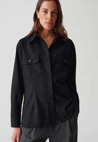Camicia nera con bottoni, realizzata in tessuto liscio, con colletto, due tasche sul petto e maniche lunghe, abbinata a pantaloni grigi con fantasie.