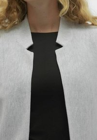 Světle šedý blazer s herringbone vzorem, s otevřeným límcem a texturovanou tkaninou, navrstvený přes černý top.