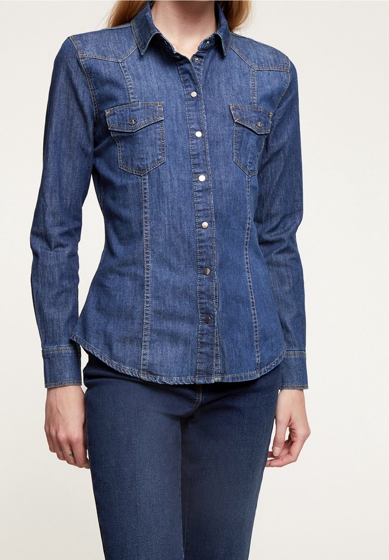 Chemise en denim foncé à boutons, manches longues, deux poches poitrine et coupe cintrée. Présente des coutures contrastantes et un col pointu.