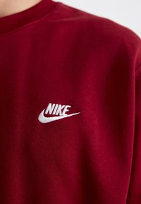 Bordeaux sweatshirt med en vit Nike-logotyp broderad på bröstet, som visar varumärkesnamnet och swoosh-symbolen.
