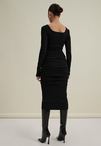 Černé přiléhavé šaty s dlouhými rukávy a čtvercovým výstřihem, se shromážděnou texturou na zádech, skombinované s lesklými černými kozačkami nad kolena.