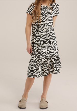 Meisje draagt een zwart-witte jurk met zebraprint en een ceintuur, gecombineerd met beige instapschoenen, staand op een lichte achtergrond.