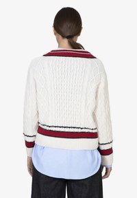Maglione lavorato a trecce color crema con colletto e orlo a coste rossi, caratterizzato da strisce navy e rosse. Indossato sopra una camicia azzurro chiaro.