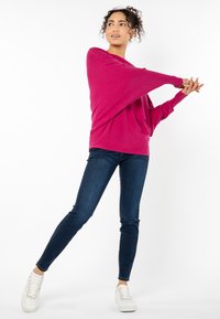 Fuchsia-übergroßer Pullover mit Fledermausärmeln, kombiniert mit dunkelblauen Skinny Jeans und weißen Sneakers. Minimalistisches Design auf einfarbigem Hintergrund.