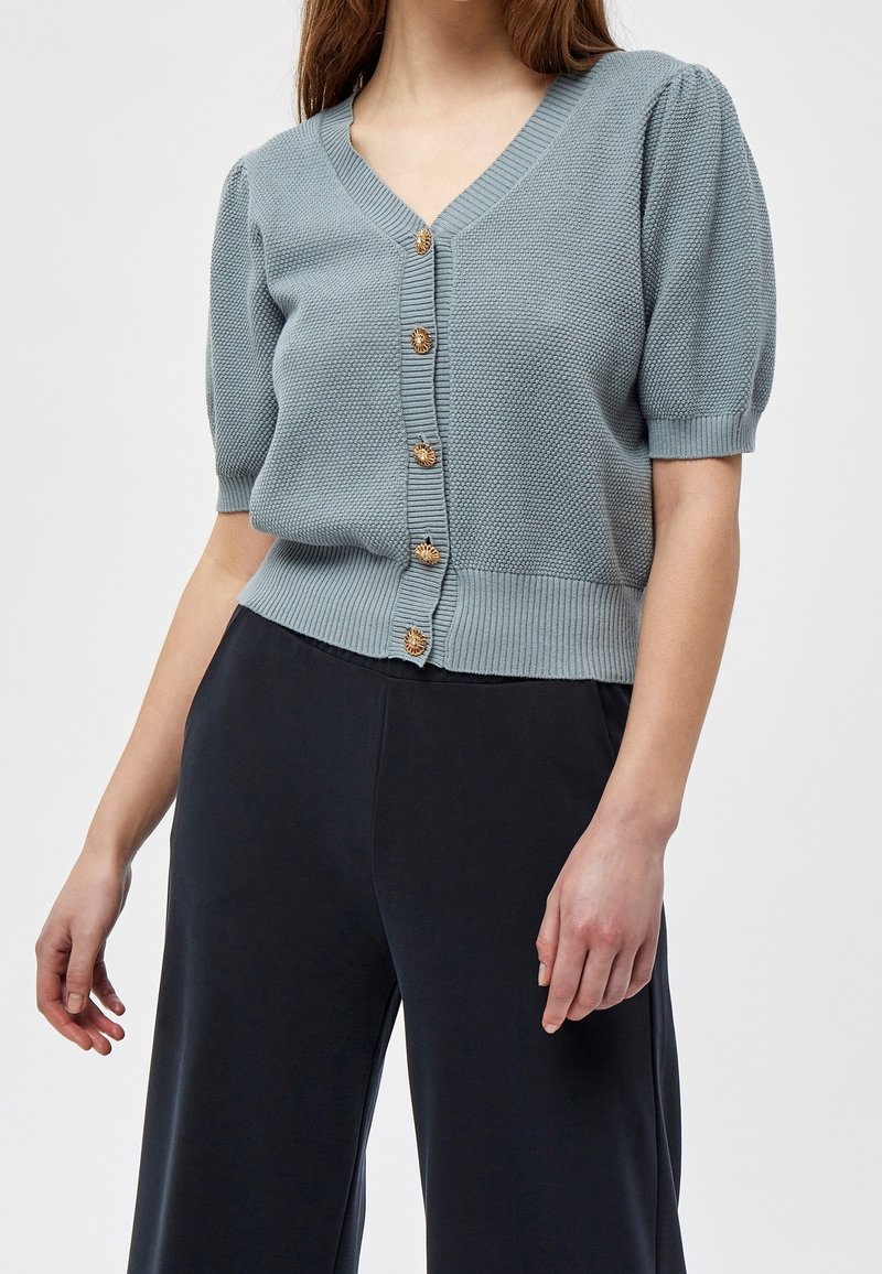 Světle modrý pletený kardigan s krátkými nabíranými rukávy, výstřihem do V, žebrovanými manžetami a lemem, s šesti kulatými zlatými knoflíky vpředu.