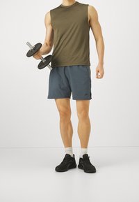 Ärmlös olivgrön linne, blå sportshorts och svarta sneakers; håller en hantel; minimalistisk design med logotyp på shortsen.