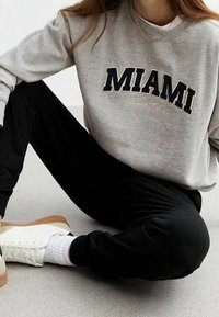 Lichtgrijze sweatshirt met "MIAMI" in marineblauw gedrukt, gecombineerd met zwarte joggerbroek en witte sneakers met een bruine zool.
