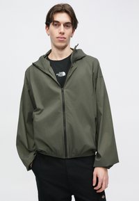Ελαφρύ χακί πράσινο μπουφάν με κουκούλα, κλείσιμο με φερμουάρ, ελαστική μέση και άνετη εφαρμογή. Φοριέται πάνω από μαύρο πουκάμισο με λογότυπο.