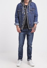 Jeansjacke mit aufgerollten Ärmeln über einem grauen Kapuzenpullover, kombiniert mit dunklen Jeans und weißen Sneakers. Merkmale sind Ziernähte und Kordeln.