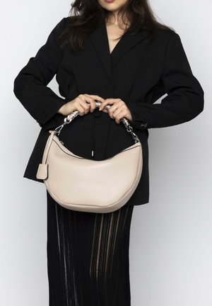 Beige leather handbag with a curved shape, silver chain handle, and smooth texture. Worn by a person in a black blazer and vertical striped skirt.