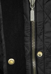 Interior de um casaco preto acolchoado com um fecho de correr prateado com a marca "Barbour", botões de pressão dourados e detalhes em tecido preto texturizado.