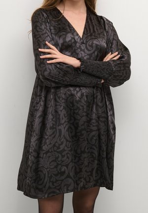 Robe noire avec un design à motifs, décolleté en V, manches longues et poignets froncés. Le tissu a une texture soyeuse avec une silhouette fluide.