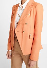 Dvouřadý blazer v jasně oranžové látce, s zlatými knoflíky, přiléhavým střihem a strukturovanými klopami. Nositý přes potištěnou košili.