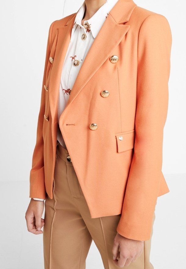 Dvouřadý blazer v jasně oranžové látce, s zlatými knoflíky, přiléhavým střihem a strukturovanými klopami. Nositý přes potištěnou košili.