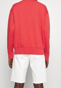 Persona che indossa un felpa rossa a maniche lunghe e degli shorts di jeans bianchi fino al ginocchio, in piedi con le mani rilassate ai lati.